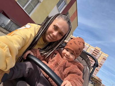 A mother and child are photographed outdoors, possibly near a residential area. The mother, wearing a yellow hoodie, is positioned at the bottom of the frame, smiling at the camera. Her hair is styled in braids with blonde and brown strands. Above her and slightly left, a young child in a brown patterned hat and a furry brown jacket is also looking at the camera and holding onto a black stroller bar. The background shows a low-rise building with a beige and red striped facade, suggesting an urban environment. The sky is visible in the upper left corner of the frame and is a clear blue. In the background, a multi-story residential building with yellow and pink accents can be seen. The specific location is not explicitly stated but is possibly in an urban area, and could be assumed to be in Yaroslavl', Russia.