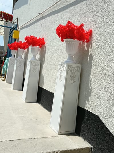The image depicts an outdoor scene in the city of Osh, Kyrgyzstan, featuring a row of four white decorative pedestals placed against a textured white wall. Each pedestal is topped with a white vase filled with bright red artificial flowers. The pedestals are rectangular and taper slightly towards the top, with decorative relief carvings on each visible face. A shadow cast by the pedestals and flowers falls on the wall. The ground in front of the pedestals is a light-colored concrete surface with visible cracks. To the left of the scene, a blue doorway or gate is partially visible, along with a yellow electrical box mounted on the wall. Further to the left, a white awning and some red floral decorations are visible above the doorway. A person wearing a headscarf is partially visible near the doorway. The overall impression is of an outdoor decoration setup, possibly for an event.
