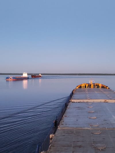 The image captures a maritime scene on a wide, calm body of water under a clear, pale blue sky, described as being near Malinukha, Russia. In the immediate foreground, the grey, flat deck of a large vessel, possibly a barge or cargo ship, is observed from a stern perspective. The deck features multiple rows of circular metal hatches and is bordered by blue railings on the port side. Towards the forward end of this vessel, a structure with yellow and orange components is visible. A discernible wake emanates from the vessel's port side.
In the midground, two additional vessels are underway, moving from left to right across the frame. The vessel positioned to the left displays a blue hull with red trim and a prominent white superstructure, indicative of a river push-boat or a self-propelled barge. To its right and slightly astern, a second, darker-hulled cargo ship or tanker, featuring a white superstructure at its stern, is also in motion. Both midground vessels are generating distinct wakes on the water.
A long, linear, dark green landmass, appearing as a distant treeline or low-lying shore, forms the horizon. The lighting suggests either dawn or dusk, with soft illumination reflecting off the calm water surface. No individuals are visible in the scene.