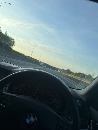 The image shows a view from inside a car, looking out through the windshield. The dashboard and steering wheel are visible, with the BMW logo prominently displayed. Through the windshield, there is a highway stretching into the distance, lined with lampposts and trees. The sky is a light blue with some wispy clouds. Based on the GPS, we are near the city of Epping Forest, United Kingdom. There are no people visible in the image.