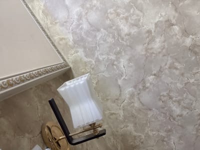 The image shows a wall with a marble-like texture and a portion of a light fixture. The wall is decorated with a marbled pattern in shades of beige and gray, with darker veins running through it. A decorative border with a gold ornamental pattern is visible along the top left edge of the image, suggesting it's part of a cornice or molding. A wall-mounted light fixture with a white, pleated lampshade is positioned on the wall, extending from the left side of the frame. The base of the light fixture appears to be made of brass or gold-toned metal and is attached to a black arm. The overall scene depicts an interior detail, likely within a building. There are no people or obvious signs of human activity in the image. The location in Shofirkon, Uzbekistan, is not visually indicated by any landmarks or contextual clues within the image itself.