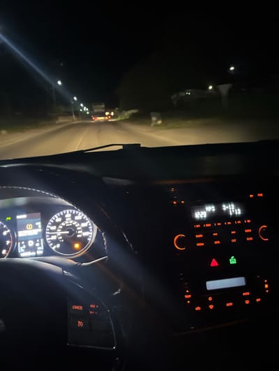 The image shows the interior of a car at night, likely in motion. The dashboard displays a lit speedometer, with the needle pointing to approximately 60. To the right, the car's stereo system is also illuminated, displaying the time as 12:29 and MP3 information. Buttons for radio, media, info, cat, and scan are visible. An "Eco Mode" indicator is lit green. Outside the car, the road is visible, illuminated by headlights and streetlights. A truck is visible in the distance, and there are buildings and trees along the side of the road. The overall lighting suggests nighttime driving conditions. The location in Arsha, Georgia is not discernible from the visual information in the image.