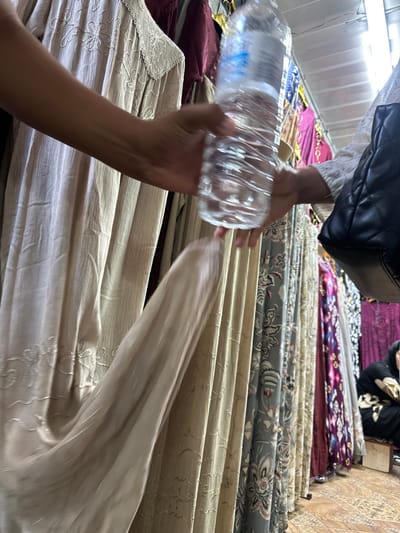 In the city of Kara-Suu, Kyrgyzstan, a person's hand holds a clear plastic bottle filled with water, its surface textured by ripples. Another person's hand, holding a black quilted bag, is visible on the right. The background features a row of colorful dresses hanging on display, with intricate patterns and varying fabrics, suggesting a market or boutique setting. The floor is tiled with a decorative pattern.