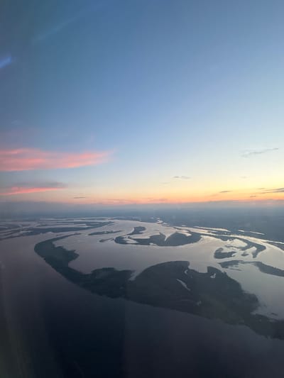 This image depicts an aerial view of a river delta or estuary at sunset. The scene is dominated by the interplay of light and shadow on the water, with a clear division between the bright, sunlit sky and the darker, shadowed landforms.
The sky transitions from a pale blue on the left to warm hues of yellow, orange, and pink near the horizon, indicating it is sunset. Wispy clouds are visible in the sky. The water itself is a prominent feature, reflecting the sky and creating intricate patterns of light and dark as it snakes through the landscape. The landmasses appear as dark shapes, possibly islands or peninsulas, bordered by the water.
The overall mood is serene and tranquil, characteristic of a sunset. There are no visible people or man-made objects that stand out. The vastness of the river system and the expansive sky suggest a natural and somewhat remote location. The specific geographical context of Kokkorevo, Russia, suggests this might be a view over the Neva River delta or a similar large river system in the region. The time of day is definitively sunset due to the colors in the sky and the low angle of the light. The weather appears clear with some scattered clouds. No text is visible in the image.