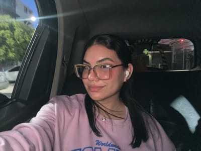 A young woman with dark hair, wearing glasses and a light pink sweatshirt, sits in the passenger seat of a car at night. She is looking at the camera with a slight smile. A white wireless earbud is visible in her right ear. A necklace with a blue and white pendant is worn around her neck. Outside the car window, trees and streetlights are visible, suggesting an urban environment. The car appears to be in motion or parked on a street in Conuco Viejo, Venezuela, as indicated by the presence of typical city infrastructure and lighting at night.