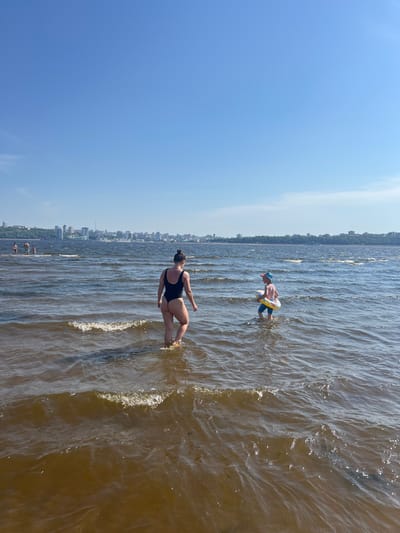 In the image, a woman in a black swimsuit walks in the water with a city skyline in the background. To her right, a young child wearing a blue cap and striped shorts, with a yellow inner tube around their waist, also walks in the water. Other people are visible in the distance, wading or swimming. The water is murky and choppy, with small waves. The sky is clear and blue. The scene takes place at a body of water, likely a river, with a densely populated city visible on the far bank. The city is identified as Sosnovka, Russia.