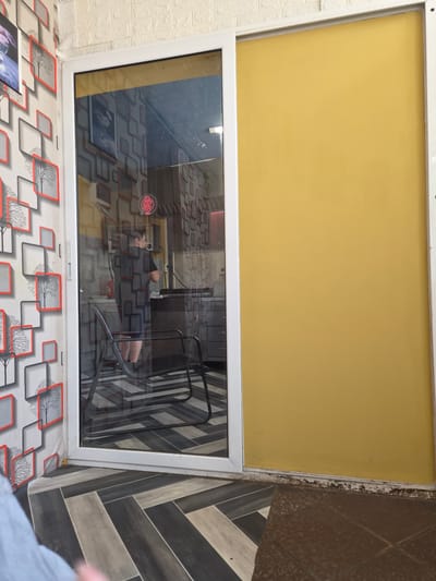 The image shows a street-level view of an establishment in Kaleytsa, Bulgaria. The scene is framed by a sliding glass door and a solid yellow panel. Inside the establishment, visible through the glass, a person is standing in what appears to be a kitchen or service area. They are wearing a dark t-shirt and shorts. The interior is decorated with a repeating pattern of grey and red squares and trees. A neon sign with Chinese characters is visible on the wall. The floor inside is tiled in a geometric pattern of black, grey, and white. Outside, the floor is also tiled, with a herringbone pattern of dark grey and lighter grey tiles. The wall to the left of the door is wallpapered with a similar pattern to the interior decoration. The bottom edge of the door frame shows some dirt and discoloration.