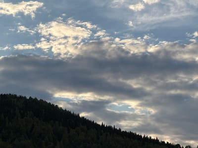 This media file captures a dramatic sky over a densely forested mountain slope, likely in Tashtagol, Russia. The scene is dominated by the sky, which is filled with a dynamic mix of clouds. Lighter, wispy clouds are illuminated by the sun, creating a bright contrast against a backdrop of darker, heavier clouds. A notable detail is a gap in the clouds, revealing a patch of blue sky.

The mountain slope is covered in a thick, dark green forest, suggesting a vast wilderness. The trees are silhouetted against the lighter sky, with their varied shapes creating an uneven, textured line along the horizon. The overall impression is one of a natural landscape under a moody, possibly late afternoon or early evening sky. The interplay of light and shadow across the clouds and the forest suggests a transition in the weather or the time of day, perhaps hinting at an approaching sunset or a break in an overcast day. No people, objects, or visible text are present in the media file. The scene evokes a sense of vastness and the raw beauty of nature.