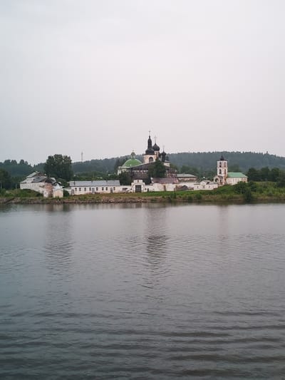 In Goritsy, Russia, a panoramic view captures the Goritsky Monastery situated on the bank of a wide river under an overcast sky. The monastery complex, an ensemble of white and light-colored buildings, features multiple domes, spires, and a bell tower. The central church boasts a prominent green dome, while other structures have darker, onion-shaped domes topped with crosses. A tall white bell tower with a dark dome stands to the right of the main complex. The buildings are arranged along a stone-lined embankment, with a narrow strip of green grass and trees separating them from the water. A dense forest forms the backdrop of the monastery. The river in the foreground is calm, with subtle ripples on its surface, reflecting the muted light. The overall atmosphere is tranquil and serene.