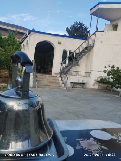 The image depicts an outdoor scene in what appears to be a courtyard or yard. In the foreground, a shiny metallic kettle is prominently displayed, positioned on a tray with a patterned surface. The kettle's reflection of the surrounding environment is visible.

The background features a building with white walls and arched entrances. A metal staircase leads up to an upper level of the building. The building has blue trim along its roofline. To the left of the building, trees are visible, and to the right, a small tree with green leaves grows. A light blue wooden bench is situated near the staircase, and another wooden table is placed further to the right. The ground is paved with concrete.

The sky is blue with scattered white clouds, suggesting a daytime setting, likely late afternoon given the timestamp. The weather appears clear and pleasant. The overall atmosphere is calm and relaxed.

Visible text at the bottom of the image includes "POCO X5 5G | EMIL BABAYEV" on the left, and the date "25.06.2026" followed by the time "18:44" on the right. This suggests the image was taken on June 25, 2026, at 6:44 PM. The location context provided is Sabunchu Raion, Azerbaijan.