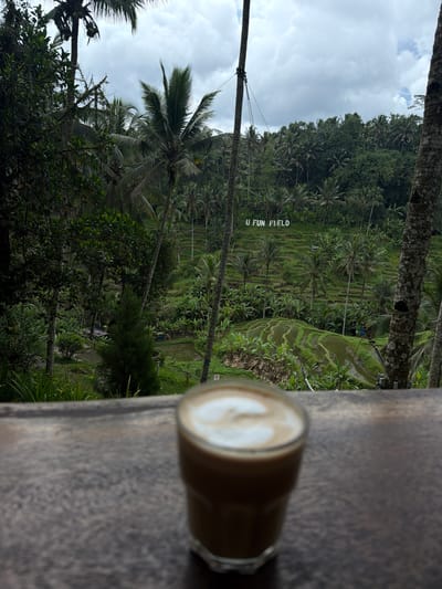 A vertical, eye-level shot shows a cappuccino in a glass in the foreground on a wooden surface. The background reveals lush rice paddies with "U FUN FIELD" spelled out in white letters on one of the paddies. Palm trees are interspersed throughout the landscape, with dense forest visible in the upper reaches of the shot. The sky is cloudy. The scene is situated in Tegal Suci, Indonesia.