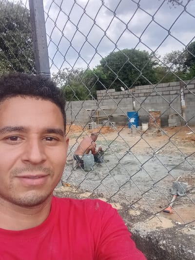 A selfie shows a man in a red shirt, partially framed by a chain-link fence. Behind the fence, two individuals appear to be engaged in construction work. A building constructed from cinder blocks is visible, and some tools sit on the ground. The background includes trees and a cloudy sky.