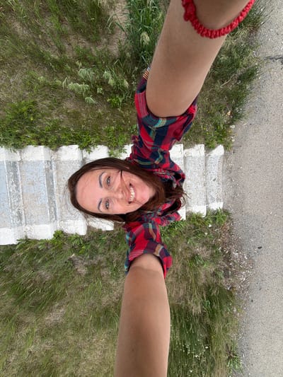 This is a high-angle, selfie-style photo taken outdoors, likely in a rural or natural setting, possibly near Nikolaevka, Russia. The subject is a woman with long, dark brown hair and blue eyes, who is smiling broadly at the camera. She is wearing a red, blue, and black plaid shirt and a red beaded bracelet on her left wrist.

The woman is positioned on a set of old, weathered concrete steps, surrounded by tall, dry grass and some scattered weeds. Above the steps, there appears to be a paved area, possibly a road or path. The overall scene suggests an overcast or moderately lit day, with no strong shadows indicating a specific time of day. The dry grass hints at either a dry season or a less fertile area.

The perspective of the photo, looking straight down at the woman as if from above, creates a unique and somewhat disorienting viewpoint. Her expression is cheerful and energetic, suggesting she is enjoying herself or the experience of taking the photo. There is no visible text in the image.