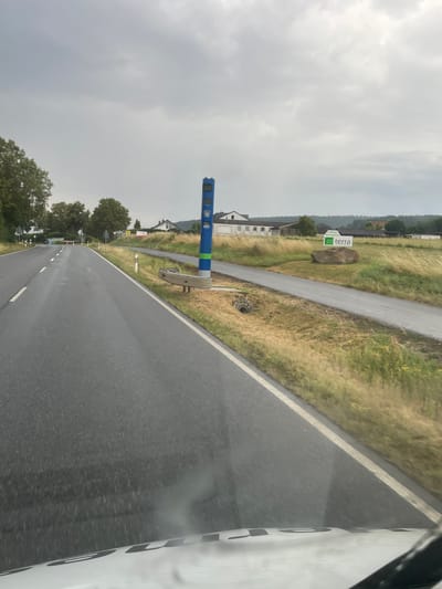 The image shows a wet, gray asphalt road in Wartenberg, Germany. The road is lined with white markers and a yellow line. On the right side of the road, there is a blue traffic camera with a green stripe. Beyond the camera is a grassy field with a large rock and a sign that reads "terra". In the background, there are buildings and trees. The sky is overcast with gray clouds. The image appears to be taken from a moving vehicle.