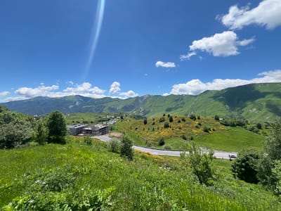 In Kumlistsikhe, Georgia, a bright, sunny day illuminates a vibrant landscape of rolling green hills and majestic mountains. The sky is a brilliant blue, dotted with fluffy white clouds and a prominent lens flare streaks across the upper left portion of the image. Below, the lush green hills are interspersed with patches of yellow wildflowers, creating a picturesque scene. A winding road cuts through the landscape, with two cars visible on it. In the mid-ground, a cluster of buildings, some under construction, suggests a developing area. Tall, mature trees dot the hillsides, adding to the natural beauty of the environment. The foreground is dominated by a verdant, grassy slope, teeming with wild vegetation.