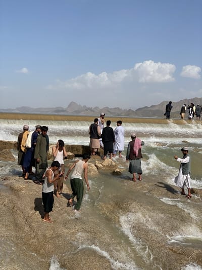 The image captures a scene at a river or waterway in Kim, Afghanistan, featuring a small dam or weir. Several men are present, some dressed in traditional Afghan clothing, including turbans and long shirts. Some of the men are wading in the water, while others stand on the dam structure. One individual appears to be using a mobile phone. The background consists of mountains and a blue sky with some clouds. The water is turbulent, with visible currents flowing over the dam. The terrain appears rocky and uneven.