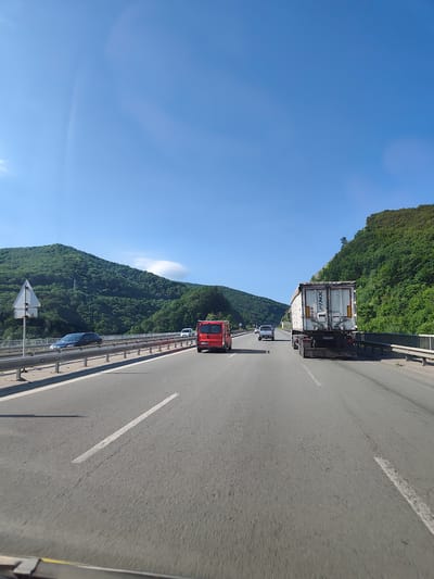 A clear, bright day with a bright blue sky is depicted, with a few wispy clouds visible. The scene is a highway passing through a mountainous, heavily forested region. The highway has multiple lanes and is bordered by guardrails on either side. Several vehicles are on the road, including a white semi-trailer truck with "P&K" branding on its trailer, a red van, and a dark blue car. The surrounding landscape is dominated by lush green mountains covered in dense trees, with some rocky outcrops visible. The perspective suggests the image was taken from inside a moving vehicle. The location is identified as being on the highway near the village of Malo Buchino, Bulgaria.
