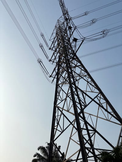 A tall, dark metal electrical tower stands against a pale blue sky. The tower is a lattice structure, with numerous crisscrossing beams and supports. Three thick, dark electrical cables extend horizontally from the tower and disappear into the sky. In the lower left and right corners, the dark green fronds of palm trees are visible, suggesting a tropical or subtropical environment. The overall impression is one of industrial infrastructure against a natural backdrop. The scene is static and lacks any human presence or activity. The location is Pakulonan Barat, Indonesia.