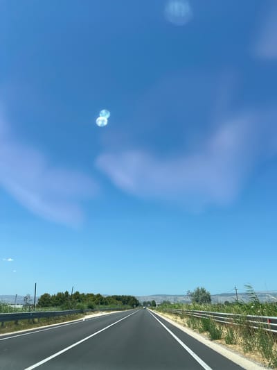 An eye-level, wide-angle shot captures a road in Manfredonia, Italy, under a clear blue sky. The asphalt road has white lane markings, leading towards a distant horizon. On either side of the road, metal guardrails line the pavement, separating it from dry, overgrown vegetation and a line of trees. The sky above is mostly clear, with a few faint, wispy clouds. Two bright, circular lens flares are visible in the upper portion of the sky, indicating the presence of sunlight. The overall scene suggests a rural or suburban setting during the daytime.
