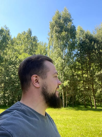 This image depicts an outdoor scene in Копнино, Russia, on a clear, sunny day. The primary subject is a male, presented in profile, facing towards the right side of the frame. He has short, dark brown hair and a full, neatly trimmed dark brown beard. His visible ear is light-skinned, and he exhibits a slight smile or smirk. He is wearing a dark grey, short-sleeved t-shirt with visible seams on the shoulder.
The background consists of a dense treeline, primarily composed of vibrant green deciduous trees (appearing like birches) mixed with darker green coniferous trees (pines) on the far right. These trees fill the middle ground, extending upwards. Above the trees, the sky is a uniform, clear light blue, indicative of a cloudless day. Below the treeline, a brightly lit expanse of well-maintained, lush green grass is visible. Strong, natural sunlight illuminates the entire scene, casting bright highlights on the foliage and grass, creating a high-contrast and vibrant appearance.