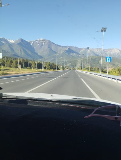 The image, taken from inside a vehicle, shows a straight, multi-lane asphalt road extending into the distance. The road is lined with streetlights, many of which are solar-powered, and a metal guardrail. On the left side of the road, there is a grassy shoulder and a line of trees. In the background, a large mountain range with snow-capped peaks rises against a clear blue sky. The scene appears to be located on the outskirts of Myk, Uzbekistan, given the presence of the mountains and the road infrastructure. The dashboard of the car is visible in the foreground, with a pair of pink sunglasses resting on it. There are no people visible in the image.