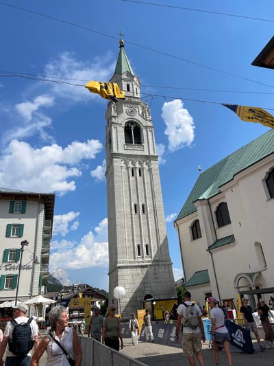This is a vertical, eye-level, outdoor photograph taken on a sunny day in Cortina d'Ampezzo, Italy. The scene is a bustling street with a prominent bell tower of a church dominating the frame. The bell tower is made of light-colored stone, with a green, pointed roof. A bright yellow flag with dark lettering is draped over the bell tower.
In the foreground and background, people are visible, some walking and others standing, suggesting a public event or tourist activity. They are dressed in casual summer clothing. Signs and banners are present, indicating a possible festival or competition. One banner, partially visible, reads "LAVAREDO TRAIL".
The weather appears to be clear and sunny, with scattered white clouds in a vibrant blue sky. The architecture of the surrounding buildings is classic European, with stone facades and shuttered windows. In the distance, a majestic, snow-capped mountain range is visible, providing a breathtaking backdrop to the town. The overall mood is lively and cheerful, with people enjoying the outdoor atmosphere. The time of day is likely midday, given the bright sunlight and clear skies.