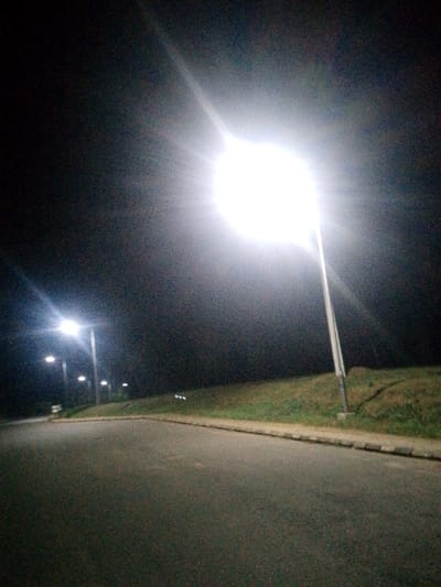 A night scene displays a paved road alongside a grassy bank. Bright streetlights illuminate the area, casting a stark glow on the road's surface. More distant lights fade into the darkness. The sky is a deep, uniform black. The grass on the bank is neatly trimmed and runs parallel to the road. The lighting creates a strong contrast between the bright, lit areas and the surrounding shadows.