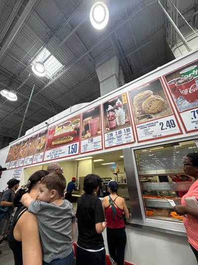 This is a medium-shot, wide-angle photograph of a Costco food court with a visible menu board and a line of customers. The environment appears to be indoors, with a high ceiling and industrial lighting. The menu board displays various food items, including hot dogs, ice cream sundaes, cookies, and smoothies, with prices and calorie information. The customers are a diverse group of people of various ages and ethnicities, some are looking at the menu, some are in line, and some are interacting with each other. A young child is being held by an adult in the foreground. Behind the counter, food service workers are visible, preparing food. The overall scene depicts a typical day at a Costco food court. There is no specific information or visual cues in the image to indicate the location is in the City of Yonkers, United States of America.