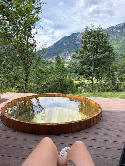 A high-angle, full outdoor shot captures a wooden hot tub on a wooden deck, set against a backdrop of lush green mountains and a cloudy sky. The hot tub, circular in shape, is filled with water that reflects the surrounding trees and sky. The deck, made of dark wood planks, encircles the hot tub, providing a clear vantage point of the tranquil scene. In the foreground, the legs of a person wearing white sneakers are visible, suggesting they are seated on the deck, enjoying the view. The mountainous landscape is dotted with trees, creating a sense of seclusion and natural beauty. The overall impression is one of peaceful relaxation in a picturesque natural setting, possibly in the vicinity of Bistrica, Montenegro.