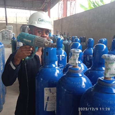 A male individual, wearing a white hard hat with a green "Gaspoint" logo and dark blue work attire, is depicted in an industrial facility in Pakuro, Nigeria. He holds a light blue heat gun with a black power cord, pointing its nozzle towards the top of a blue gas cylinder positioned directly in front of him. Numerous identical blue gas cylinders, many equipped with white valve caps bearing the "Gaspoint" logo and white labels displaying technical specifications including Chinese characters and pressure ratings (e.g., "200BAR"), are arranged in rows filling the foreground, mid-ground, and background. The environment features a concrete floor, grey walls, metal structural beams, and visible green foliage in the upper right background. A digital timestamp "2025/12/9 11:28" is present in the lower right corner of the image.