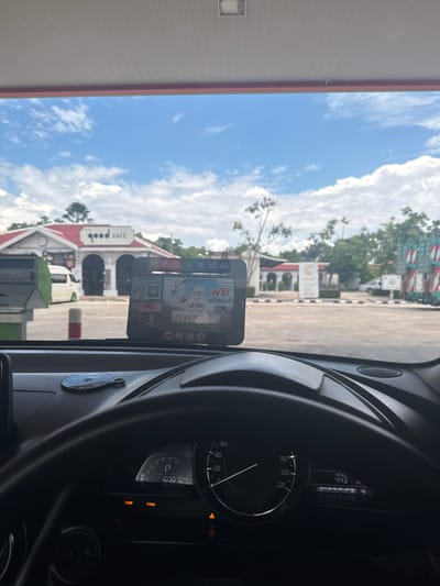 The image shows the interior of a car, looking forward through the windshield. The car appears to be at a gas station, with a "good CAFE" sign visible on a building in the distance. The sky is blue with white clouds, suggesting it is daytime and the weather is fair.

Inside the car, the dashboard is visible, displaying the car's range as 443 km and an odometer reading of 103070 km. There is also a speedometer and a tachometer. A small black device, possibly a GPS or dashcam, is mounted on the windshield.

Text visible includes "good CAFE", "FAST 3", and "ฟรี! แก้วแรงใจ" which is Thai for "Free! Glass of Heart". The location context of Ban Yid, Thailand, indicates that this is likely a gas station and cafe in that region. There are no people clearly visible in the image.