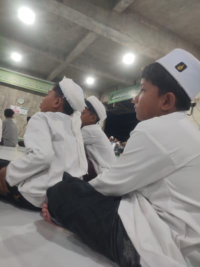 The photo shows three boys sitting on the floor inside what appears to be a mosque in Meuriya, Indonesia. They are all dressed in white shirts and black pants; two wear white turbans, while the boy in the foreground wears a white peci cap with a small logo. They are looking towards the front of the mosque. Other individuals are visible in the background, also sitting. The mosque's interior has a concrete ceiling with recessed lighting. Arabic script is visible on the wall behind the boys.