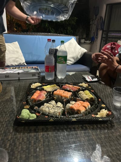 An outdoor evening scene is depicted on a residential patio in Yehud Monosson, Israel. A dark, woven-patterned glass-top table occupies the foreground, displaying a large black compartmentalized sushi platter. The platter holds multiple varieties of sushi rolls, including those with salmon, tuna, and rice coated in black sesame seeds, alongside portions of green wasabi and pickled ginger.

Behind the sushi platter, three plastic beverage bottles stand on the table: a clear water bottle, a dark soda bottle, and a Sprite bottle. To the left, a silver metallic case labeled "POKER" is visible.

Two individuals are partially visible:
1.  On the left, a person wearing a light top and khaki shorts is seen from the chest down, holding a transparent plastic dome lid above the table.
2.  On the right, another person is partially visible from the chest down, with hands holding a red, spherical, textured object. This person wears a ring on the left hand and a bracelet on the right wrist. A transparent plastic cup containing ice and liquid is also present on this side of the table.

In the background, a blue cushioned outdoor sofa with white pillows is positioned against a dark backdrop of foliage and residential elements, illuminated by artificial light, indicating a nighttime setting.