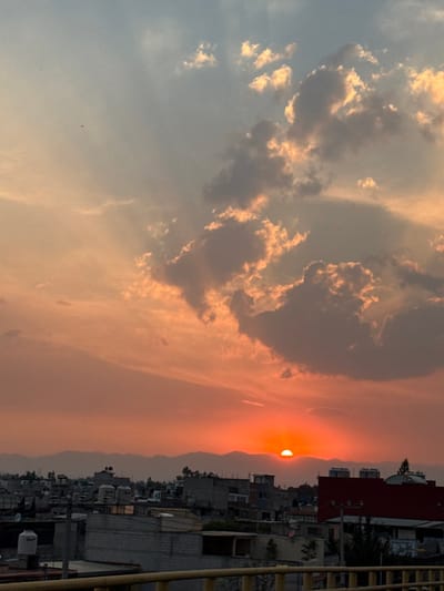 A panoramic photograph depicts a sunset over the city of Nezahualcóyotl, Mexico. The prominent sun, a vibrant orange-red semicircle, is visible near the center-right of the lower horizon, partially obscured by a distant, silhouetted mountain range. The sky transitions from deep orange and red tones near the horizon to lighter blue-grey at the top. Scattered clouds, illuminated by the setting sun, display shades of orange, grey, and dark grey, with discernible crepuscular rays radiating from behind them. In the upper left quadrant of the sky, several small, dark specks, consistent with distant birds or kites, are visible.
Below the horizon, a dense urban landscape unfolds, primarily consisting of low-to-mid-rise buildings with flat roofs, characteristic of residential or mixed-use structures. Many rooftops feature white or light-colored cylindrical water tanks. Sparse green vegetation is interspersed throughout the built environment. A distinctive building with a prominent red roof is visible on the right side of the urban sprawl. In the immediate foreground, a yellow metal railing is present, indicating the viewpoint is from an elevated position. No individuals, specific actions, or interactions are discernible within the frame.