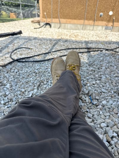 A first-person perspective shows a person sitting with their legs crossed on a bed of light gray gravel. They are wearing dark gray pants and tan work boots with yellow laces. Behind the person, a large, tan, rectangular structure with vertical ribbing is visible, supported by a concrete base. Several black cables are strewn across the gravel in the foreground and midground. In the background, a chain-link fence encloses an area with trees and what appears to be a small, wooden structure. The scene is outdoors and appears to be an industrial or utility area, possibly in Des Moines, USA. The lighting suggests it is daytime.