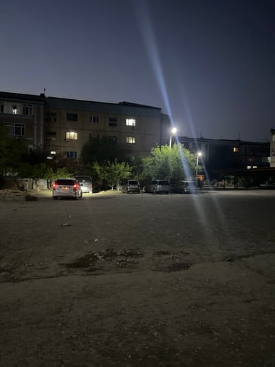 The image shows a nighttime street scene in the city of Şamaldı-Say, Kyrgyzstan. A multi-story apartment building with lit windows is visible on the left side of the frame. In front of the building, a paved area serves as a parking lot with several cars. One car, a silver hatchback, is parked in the foreground, its taillights illuminated. Other vehicles, including a light-colored minivan and a darker sedan, are parked further back. Trees are scattered throughout the scene, providing some greenery against the urban backdrop. Streetlights cast long beams of light, illuminating the ground and creating strong highlights. The sky is dark, suggesting late evening or early morning. The overall environment appears to be a residential area with a mix of concrete and gravel surfaces. Two individuals are seated near a large rock on the far left, partially obscured by trees.