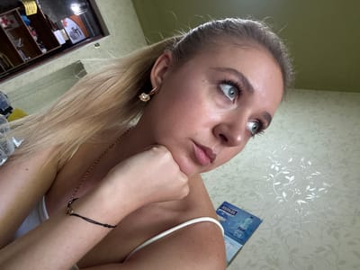 A young woman with blonde hair pulled into a ponytail is shown in a close-up shot, looking off to the right of the frame. Her left hand is resting on her chin, with her fingers curled. She is wearing a white tank top and a gold chain necklace, as well as a bracelet with a black string and a small heart charm. Her right earlobe has a gold earring with a clear stone. The background consists of a patterned wallpaper and a shelf unit with various items. The scene is lit by artificial light.