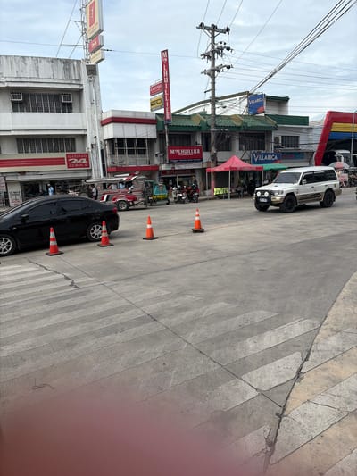 The image captures a street scene in Dagupan, Philippines, likely near a commercial area. In the foreground, a pedestrian crossing is visible on a concrete road. Several orange traffic cones are placed on the road, some near the pedestrian crossing and others in the middle of the lane.
A black sedan is parked on the left side of the road, positioned near a pedestrian crossing. To the right of the sedan, a multi-colored jeepney is on the road, occupied by passengers. Further down the road, a white SUV with "Mitsubishi Pajero" branding is parked.
Various businesses are visible along the street. Signs for "M LHUILLIER," "GISING 24 ORAS," and "VILLARICA" are prominent on the buildings. Tents, possibly for vendors, are set up in front of some establishments. The sky is overcast with visible utility poles and power lines. The overall impression is that of a typical urban street environment in the Philippines.