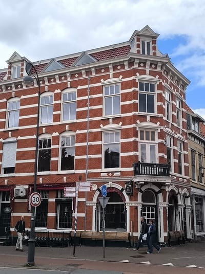 The image depicts a corner building in Haarlem, Netherlands, constructed with red brick and white horizontal banding. The building features numerous windows of various sizes and designs, including arched windows and a balcony. The architecture suggests it is a historic structure.
In the foreground, a street scene unfolds. A man in a light-colored jacket and pants is walking away from the camera, holding a bag. He is near a lamppost with a sign displaying "30" within a red circle, indicating a speed limit. Across the street, a couple is walking towards the camera. The woman is wearing a dark jacket and pants, while the man is in a darker jacket and pants. They are walking on a sidewalk in front of the building.
Several street signs are visible, including signs indicating "Alkmaar," "Zaandvoort," and "Amsterdam," suggesting these are directions to different locations. A bicycle symbol is also present on a blue sign. Benches are placed along the sidewalk in front of the building, and there are awnings over some of the ground-floor windows. The sky is partly cloudy, with patches of blue visible. The overall environment is urban, with the building occupying a prominent corner.