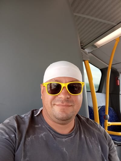 A man is taking a selfie inside a public bus, possibly in Zofingen, Switzerland. He is wearing a white skullcap and bright yellow sunglasses, which reflect a street scene. He has short, dark stubble and is smiling faintly. He is wearing a dark grey t-shirt with some lighter, faded markings on it. A thin white string from his shirt is visible near his neck. Yellow poles are visible on either side of him, characteristic of bus interiors, and blue and red patterned seats are in the background. The bus appears to be well-lit by artificial light, suggesting it could be daytime or evening. The overall atmosphere is casual and relaxed, with the man appearing comfortable and perhaps amused.