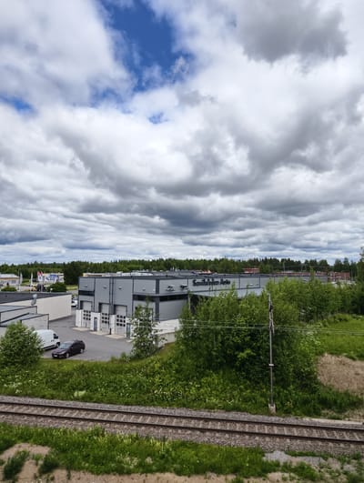 The image depicts a grey, modern, industrial building with the text "Savilahti Auto" visible on its facade. The building has multiple large garage doors. Adjacent to it are smaller buildings and a paved area with a white van and a dark-colored car parked. The building and its surroundings are framed by lush green trees and vegetation. In the foreground, a set of train tracks runs horizontally across the lower portion of the image, bordered by more green grass and some dirt. The sky above is filled with dramatic, dark grey clouds, with patches of blue sky peeking through. The overall scene suggests a commercial or industrial area in Mikkeli, Finland, possibly a car dealership or repair shop, under a cloudy sky.