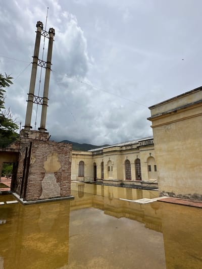 The image depicts the exterior of an old building with a reflection pool in the foreground. The building appears to be constructed of yellow stucco and features arched windows. A prominent brick structure with three tall, weathered chimneys stands to the left of the building. The chimneys are connected by metal supports. The reflection pool is filled with murky, brownish water, reflecting the building and the chimneys. The sky is overcast with dramatic clouds. In the distance, mountains can be seen. The overall scene suggests an abandoned or historic site, possibly part of the old paper mill in San Agustín Etla, Mexico. There are no people visible in the image.