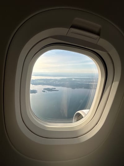 This image is an aerial view from an airplane window, looking out over a body of water with several islands and a shoreline. The water is calm and reflects the sky, which is partly cloudy with a clear horizon. The landscape appears to be a mix of green terrain, possibly fields or forests, and some structures that suggest human habitation. The window frame is a typical airplane window with multiple layers and a small latch mechanism visible at the top. The lighting suggests it is daytime, possibly late afternoon due to the angle of the sun. Without further information or contextual clues, it is impossible to definitively identify the exact location within Notre-Dame-de-l'Île-Perrot, Canada, as the image only provides a general aerial perspective of a waterscape with islands.