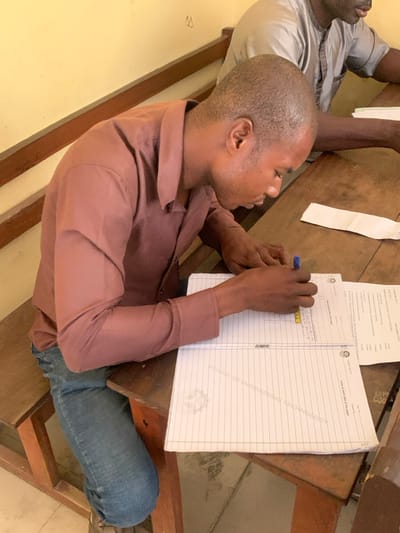 The image depicts an indoor scene within a room in Abobo, Nigeria, featuring two males seated at wooden desks. In the foreground, a dark-skinned male with short hair, wearing a brown long-sleeved collared shirt and blue jeans, is bent forward, actively writing on a lined paper with a blue pen in his right hand. His left hand rests on the paper. A small yellow ruler or straight edge is positioned on the paper near his right hand. The paper displays the watermark "SCHOOL OF ENGINEERING TECHNOLOGY" and contains visible text, including numerical entries. To the right of the central figure's workspace, a folded white paper is present on the dark wooden desk. In the background, to the right, a second dark-skinned male is partially visible, wearing a light grey textured short-sleeved top, also seated at a wooden desk with papers. The wall behind the individuals is painted a light yellow or beige, with a dark wooden horizontal rail running across it. A faint, inverted "W" or "M" symbol is marked on the upper left portion of the wall. The furniture consists of simple wooden desks and benches, characteristic of a classroom or examination hall.