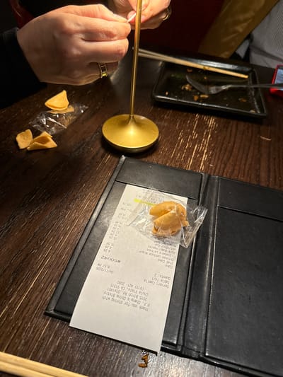 A restaurant scene in Chula Vista, California, United States of America shows a table with a black bill holder on a wooden surface. The bill holder contains a bill from P.F. Chang's China Bistro at 2015 Birch Rd., Ste 1401, Chula Vista, CA 91915, showing a date of 04/11/2026 and a check number #60042. Next to the bill is a sealed fortune cookie in a plastic bag. A person with a gold ring on their finger is holding a chopstick. Next to their hand are open fortune cookies in transparent plastic. There is also a nearly finished plate of food with chopsticks beside it.