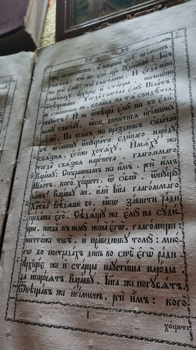 The image is a close-up shot of an open, aged book with handwritten text. The book appears to be of historical or religious significance, possibly a manuscript or an old printed edition, judging by the script and paper quality. The text is in an archaic Slavic language, likely Church Slavonic or an early form of Ukrainian or Russian, written with a quill or similar writing instrument. The ink is dark and has slightly bled into the paper. The page is yellowed and shows signs of wear, with some creases and possibly minor water damage. The writing is dense and fills the majority of the page, arranged in lines. There is also decorative border work along the edges of the page, composed of a repeating pattern of loops. In the background, out of focus, is a glimpse of a room with what appears to be a dark wooden piece of furniture and possibly a framed picture or icon on the wall. The overall impression is one of antiquity and a preserved piece of history. The location of Vyshka, Ukraine, is not visually identifiable from the image itself; it is external information provided with the prompt.