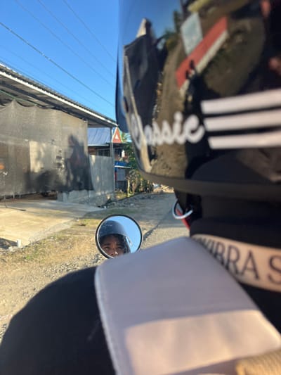 A close-up, first-person perspective shot shows a person wearing a black motorcycle helmet with white stripes and the word "Classic" in cursive on the back. The person's face is visible in a small, round rearview mirror mounted on the handlebars, reflecting their eyes and a portion of their helmet. The background depicts a street scene in Welado, Indonesia, with a building made of concrete blocks and a corrugated metal roof. A black mesh net is draped over the building's exterior. The ground is a mixture of dirt and gravel. In the distance, several houses with blue and white roofs are visible, along with power lines stretching across a clear blue sky. A triangular traffic sign with a red border and white interior is also present. The sunlight casts long shadows, suggesting it is late afternoon or early morning. A dark shadow of the rider and their vehicle is cast on the ground. A beige strap with dark lettering is visible on the back of the rider's jacket.
