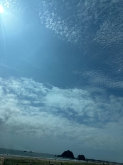 A wide-angle, slightly tilted, overexposed outdoor shot captures a bright, partly cloudy sky over a beach. The sun, located in the upper left corner, creates a lens flare and casts a bright light across the upper portion of the frame. The sky transitions from a pale blue at the bottom to a deeper blue at the top. Scattered clouds are present throughout the sky, with some appearing in wispy formations and others in more defined, textured patterns.
In the lower portion of the image, the ocean meets a sandy beach lined with sparse green grass. Two dark, rocky islets are visible in the water, with the larger one to the left and a smaller one to the right. A lighthouse, white with a dark top, stands on a rocky outcrop in the distance, to the left of the islets. A few indistinct figures can be seen walking on the beach and in the shallow water. The overall impression is a serene, coastal landscape bathed in bright daylight.