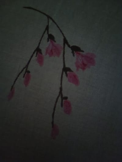 A dark, grainy image depicts a close-up of a painted or embroidered branch with pink flowers against a greyish-green background. The branch is thin and dark, curving from the upper left towards the center and bottom of the frame. Several pink, somewhat abstract floral shapes are attached to the branch at various points. The flowers appear to be in different stages of bloom, with some more tightly formed and others more open. The background has a textured appearance, suggesting fabric or a painted canvas. The lighting is dim, casting the scene in shadow. The image does not provide any information about the city of Ochiul Alb, Moldova.