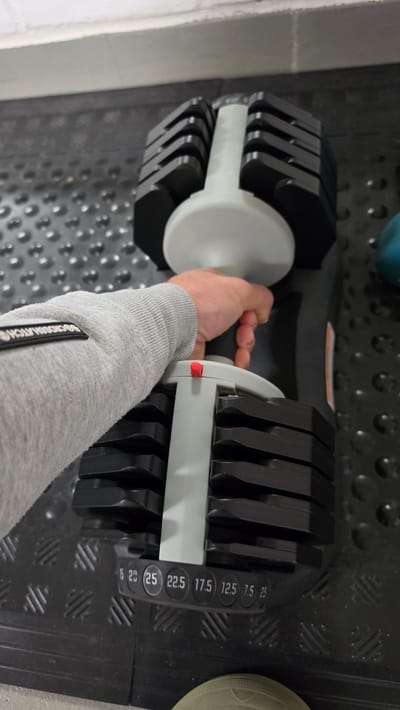 The image shows a person's arm and hand holding the handle of an adjustable dumbbell in a weight rack. The dumbbell is black and grey with weight markings in kilograms on the side. The person is wearing a grey long-sleeved shirt. The rack appears to be on a black rubber mat. The location, Ihringshausen, Germany, is not discernible from the visual information.
